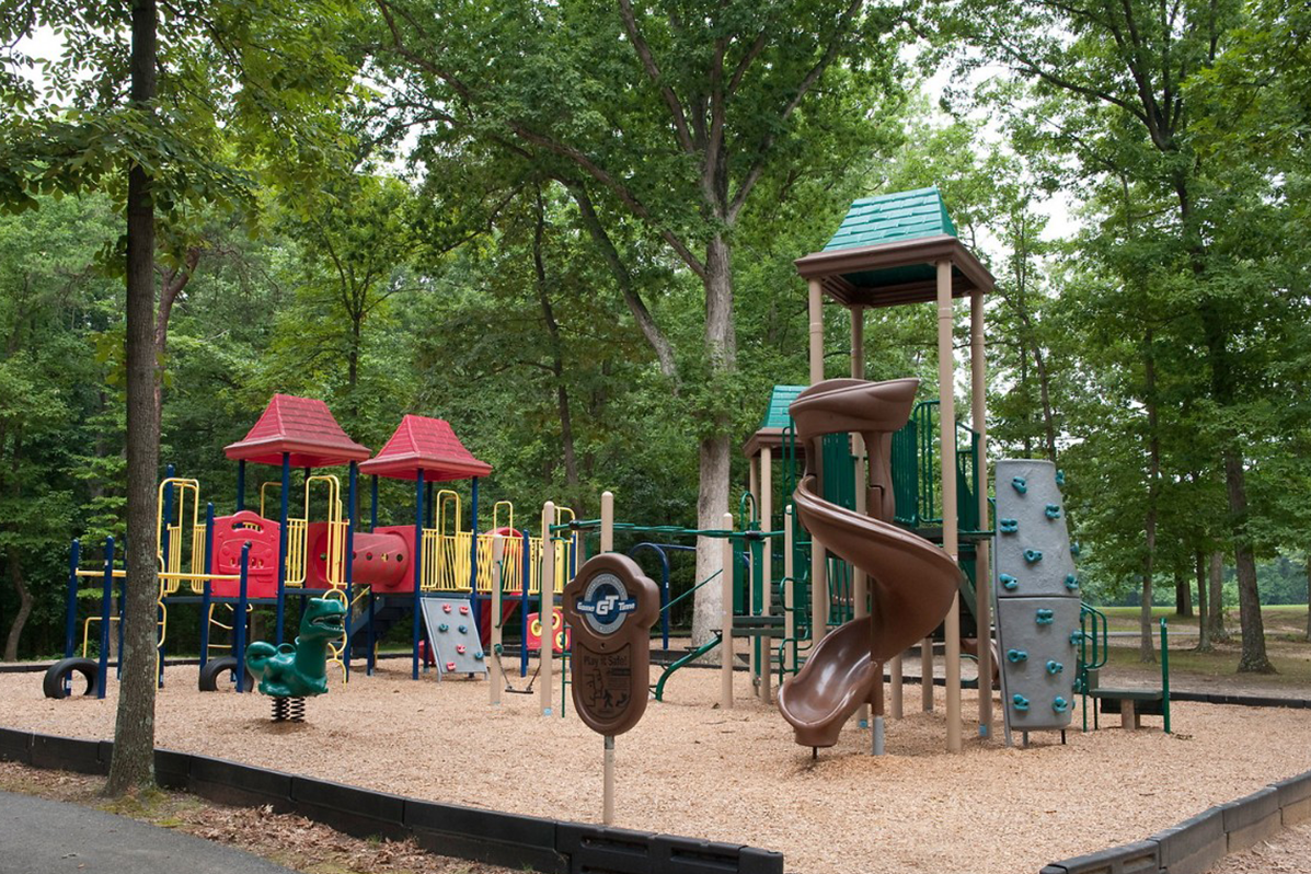 An outdoor playground with several play structures featuring slides, climbing walls, raised platforms, rockers, and covered central towers. Tall trees surround the playground on several sides.