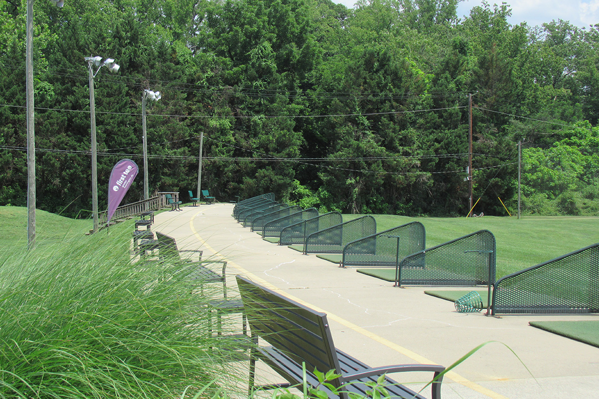 The hitting area of an outdoor driving range, with a dozen turf pads in a long row separated by low screens. Benches and chairs are nearby.