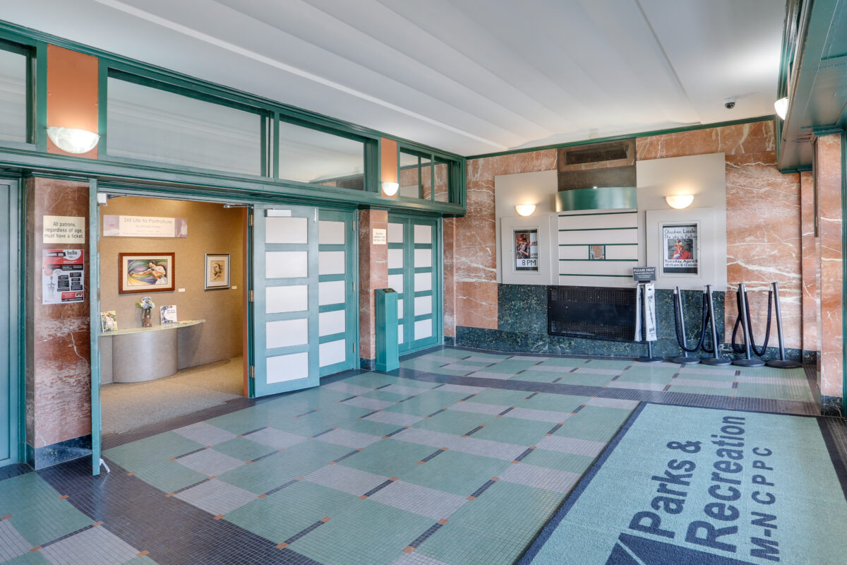A lobby with reddish marble walls and decorative tile flooring leading to a bank of green, Art Deco doors. Just inside the exterior doors is a large, green doormat with the Parks and Rec logo on it.
