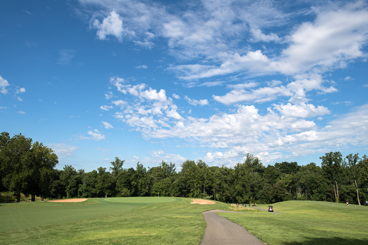 A paved path with golf carts on it winds through a grassy field. Two sand traps are visible in the far distance.