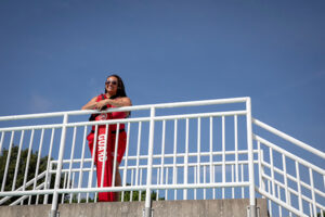A smiling lifeguard wearing sunglasses and red clothing stands outside leaning on a white handrailing.