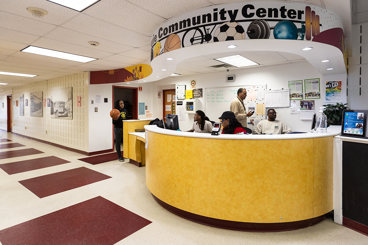 A reception desk with a curved gold wall and a sports-themed mural above reading SOUTH BOWIE COMMUNITY CENTER. To the left is a hallway with burgundy and beige tiles.