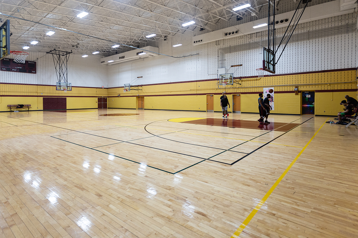 A gymnasium with a wooden floor and multiple basketball goals. Several adults stand near one basket. The room decor is gold and burgundy.