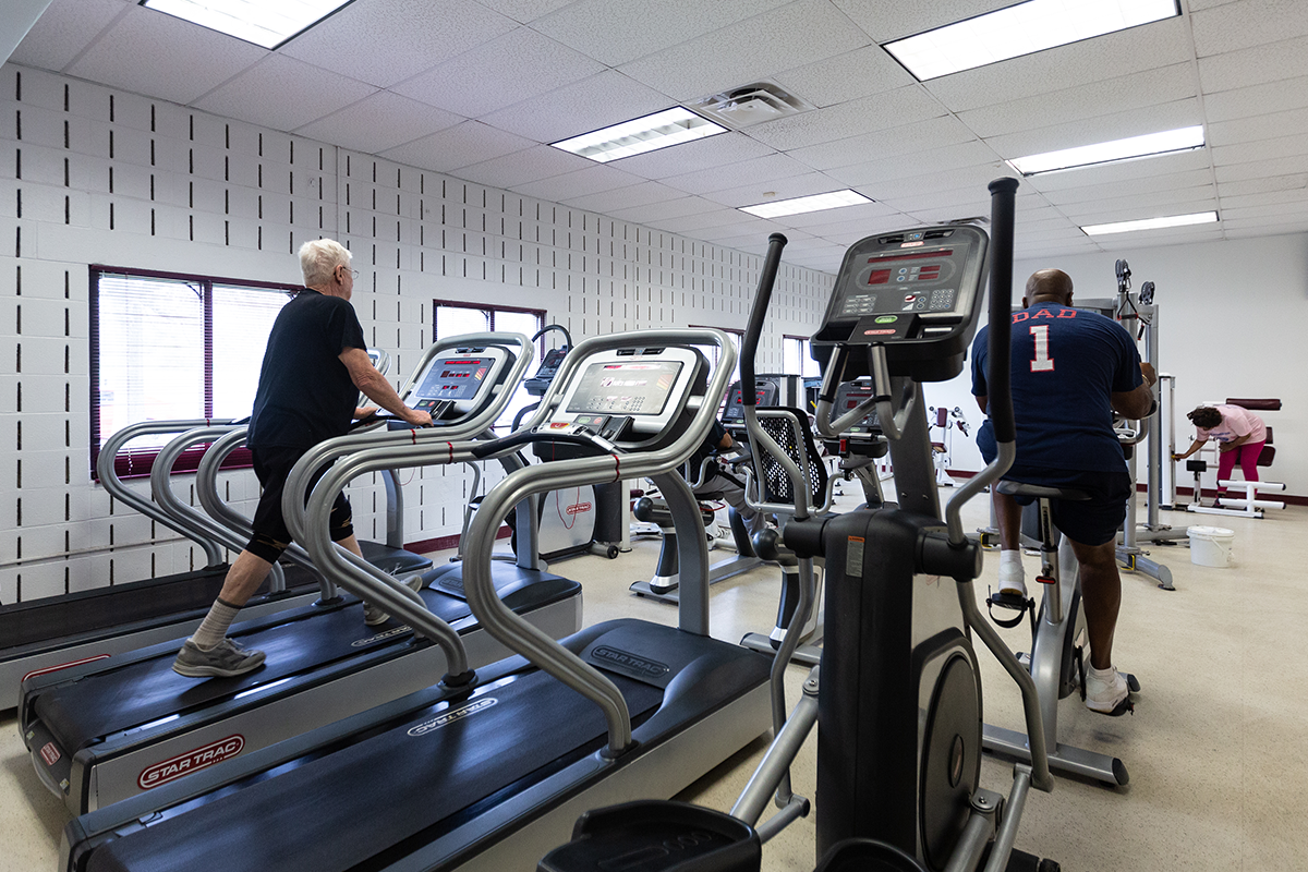 An indoor fitness room with treadmills, stationary bikes, and weight machines. There are several windows in the far wall. Two patrons are using a treadmill and a stationary bike.