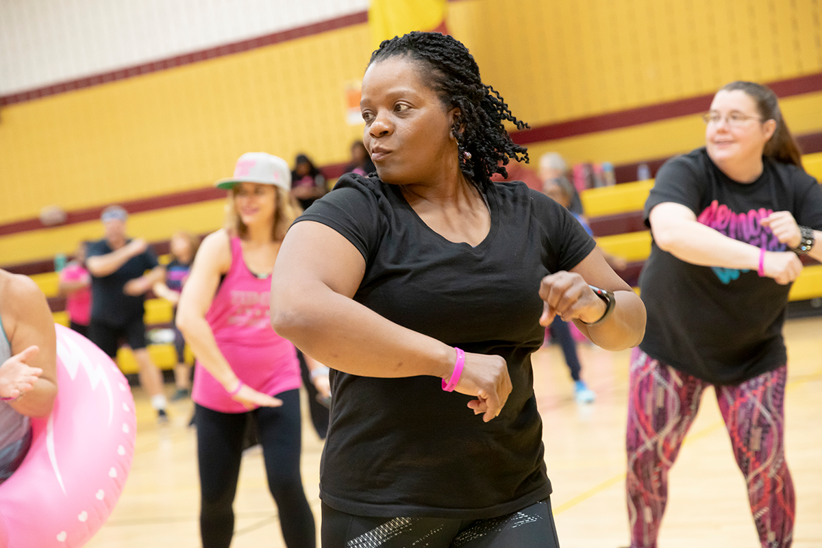 Several women in exercise clothing dance in a large gymnasium with burgundy and gold walls.
