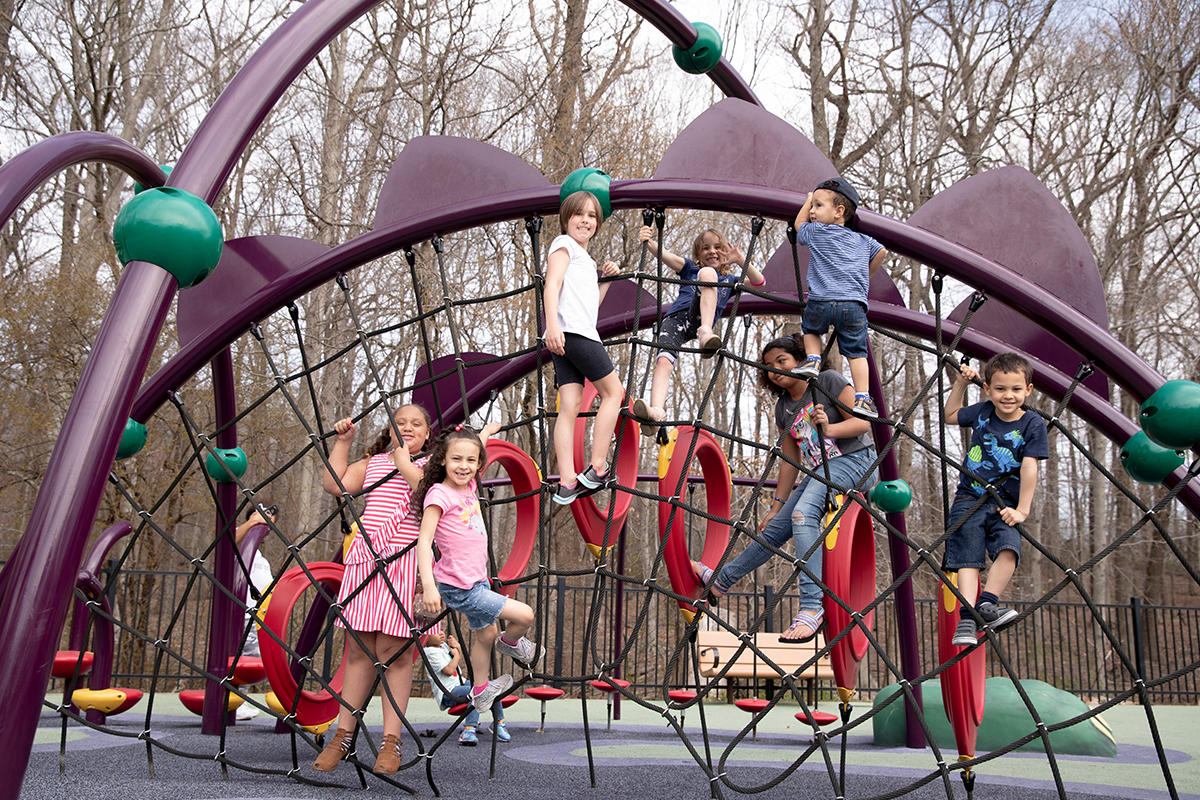 Seven smiling children climb on a large, spiderweb-like rope feature at an outdoor playground. Behind them are other climbing rings and stepping stone obstacles. There are trees in the distance.