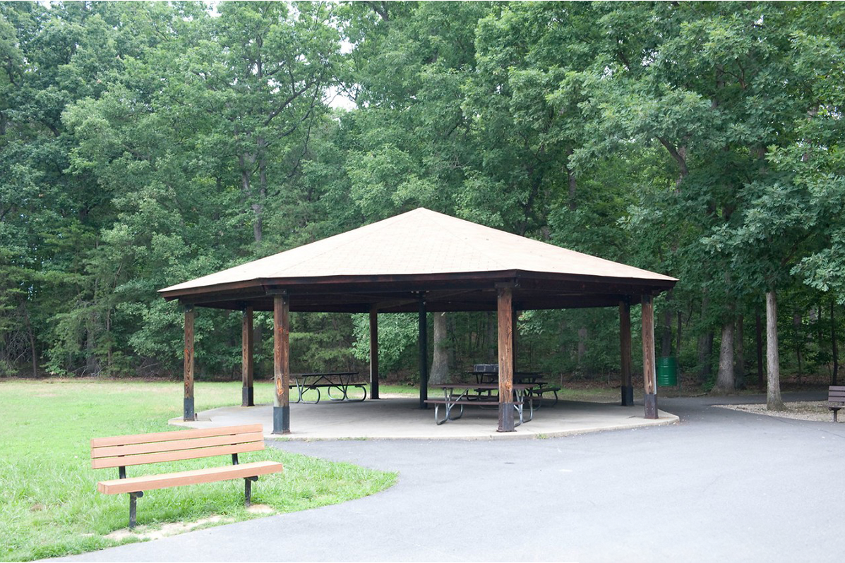 An octagonal picnic shelter covering several picnic tables on a concrete patio. An asphalt surface surrounds about half the shelter; grass and trees surround the far half.