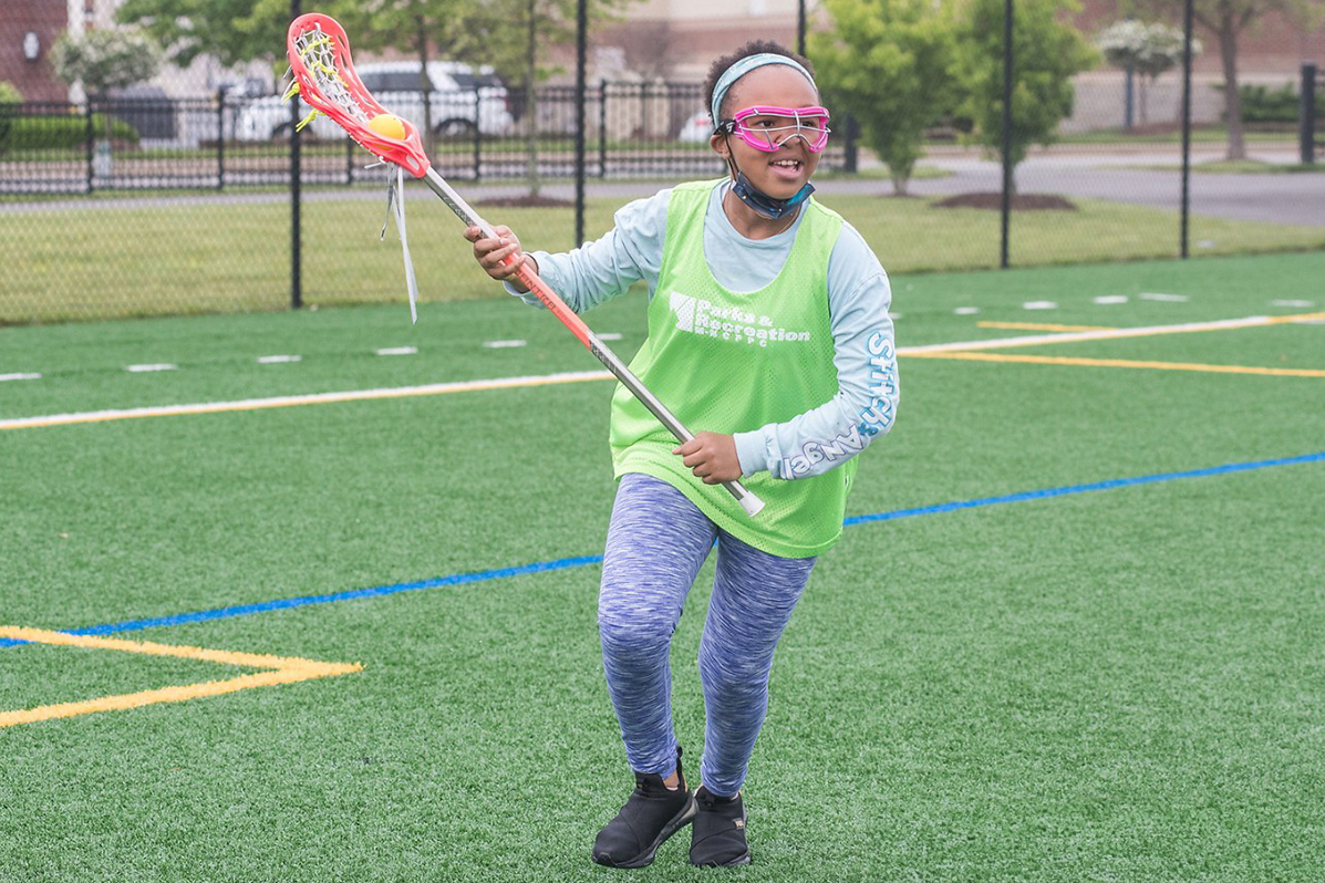 A child in hot pink glasses runs on a green field while holding a lacrosse stick.