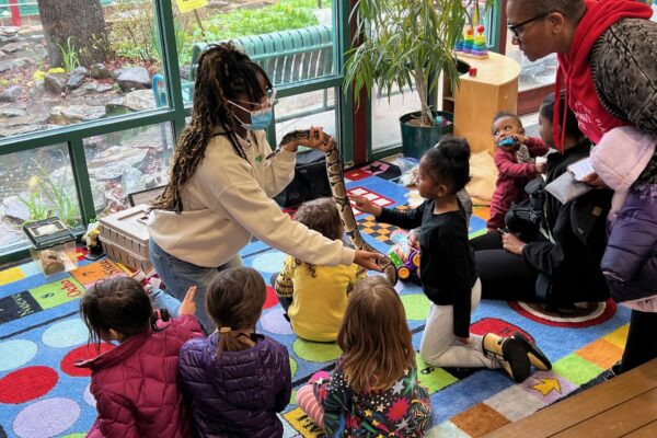 A staff member holds out a large snake to a group of seated children on a colorful rug. One child feels the snake's skin.