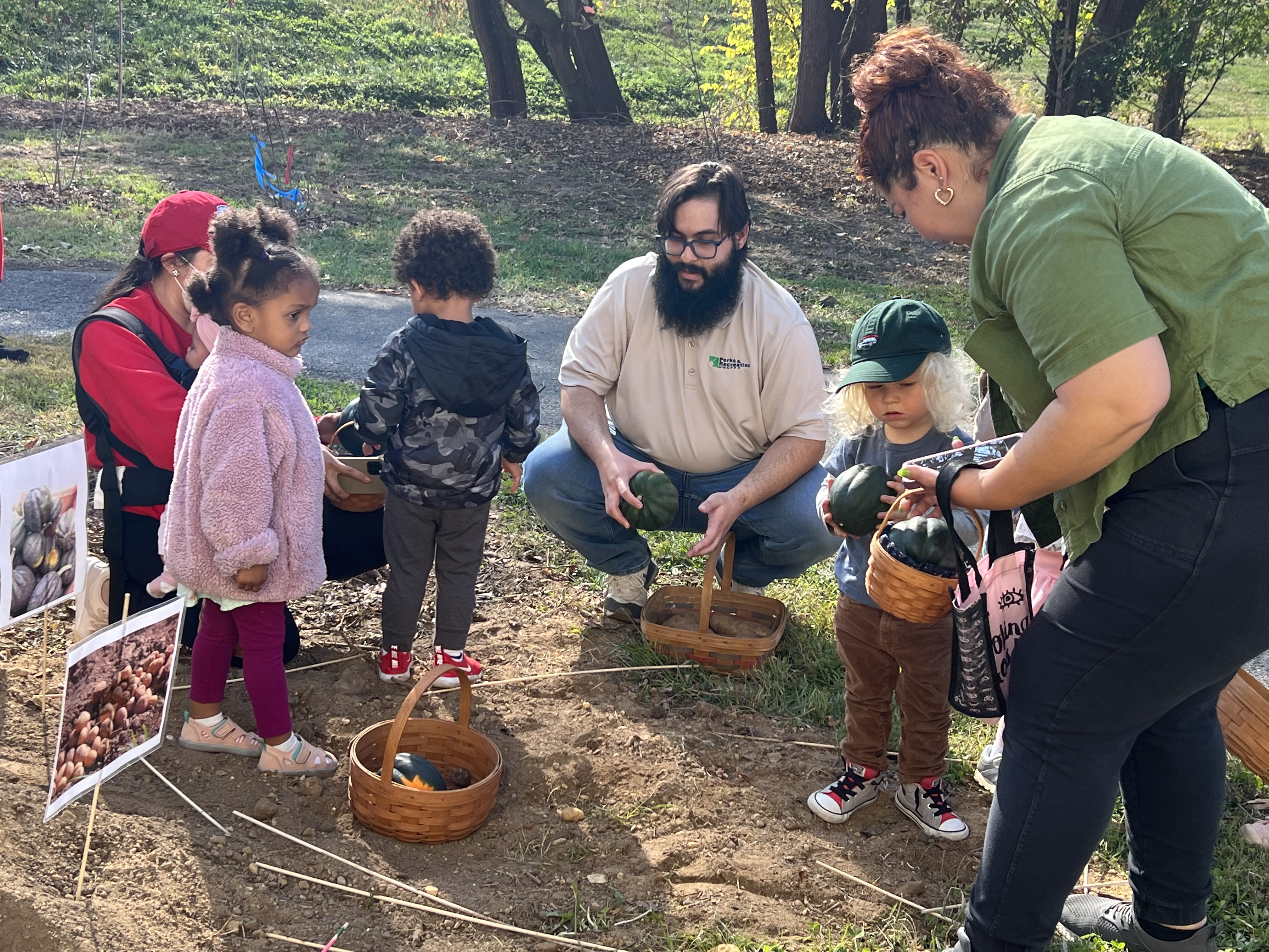 Several toddlers and adults gather on a dirt patch in the grass. Some hold baskets of acorn squash and potatoes. Signs stuck in the dirt have images of squash and potatoes.