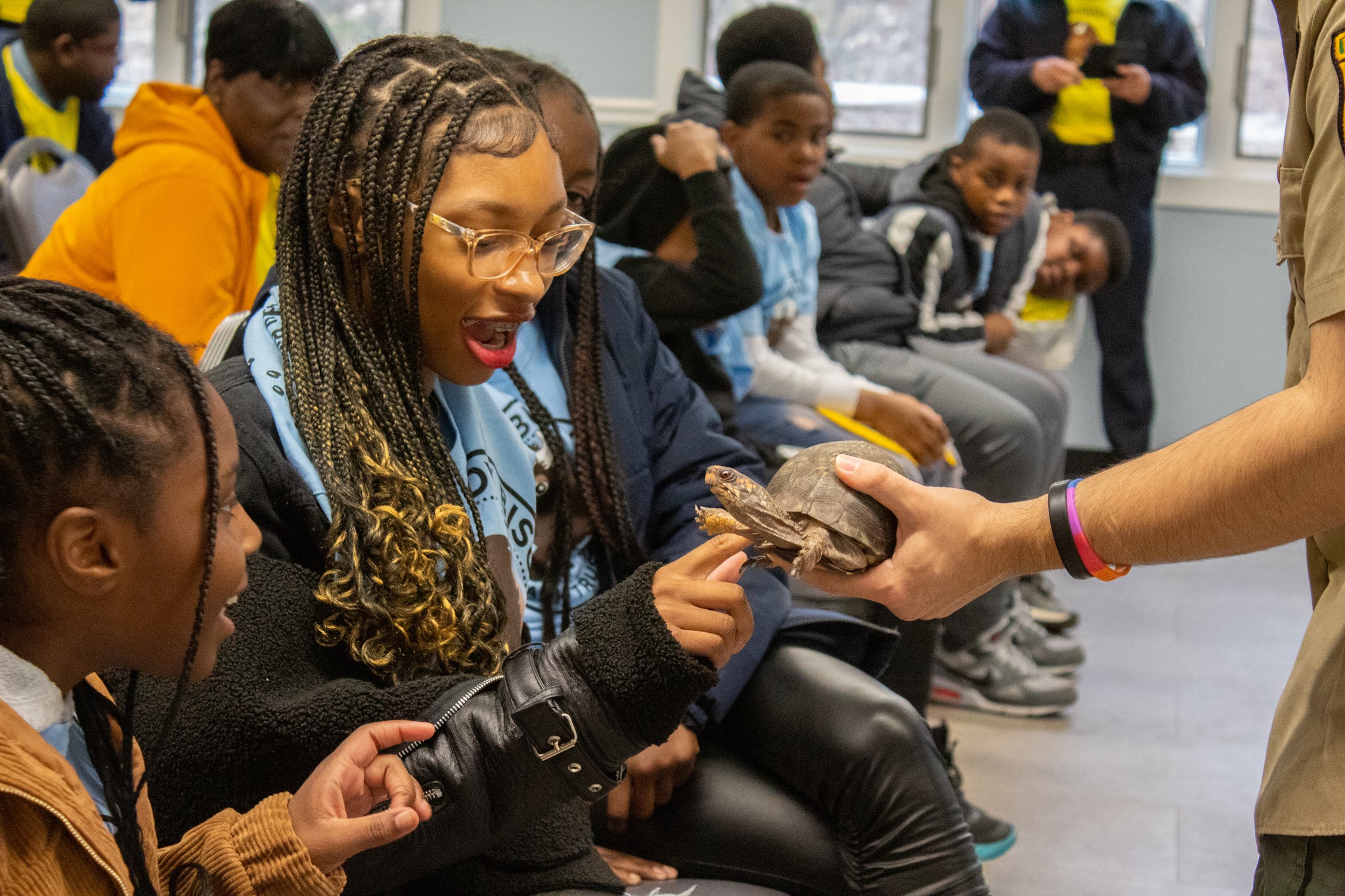 An unseen ranger holds a turtle out to a group of teens sitting in front of him. A girl with glasses and braids touches the turtle with her finger while her classmates watch.
