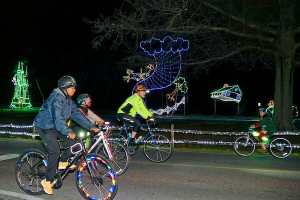 Two adults and a youth riding bicycles are passing holiday light decorations shaped like the Emerald City, a tornado, and a house flying through the air.