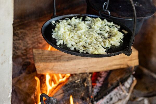 Diced onions cook on an iron plate with a curved rim, like an upside-down frisbee, that is hanging from a curved handle over an open, woodburning fire indoors.