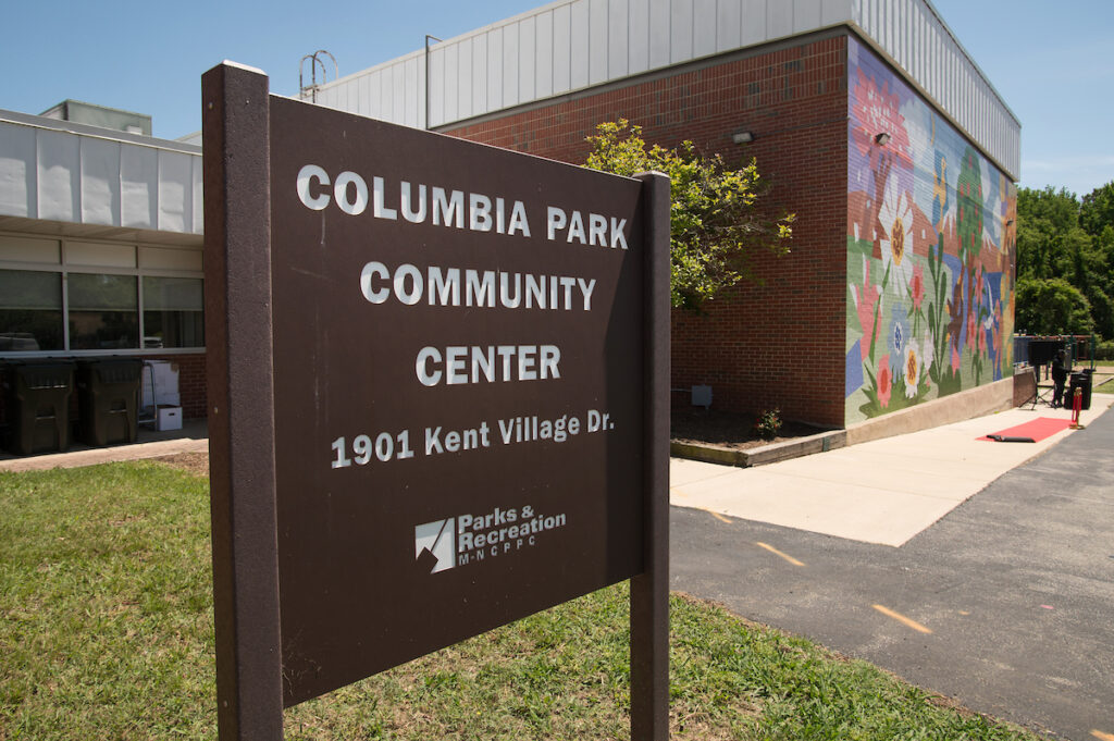 A brown sign reading Columbia Park Community Center in the grass in front of a large, brick building. One wall of the building is covered by a colorful mural.