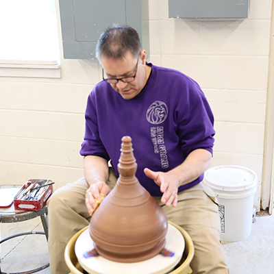 A man in glasses and a purple shirt shaping clay into a shape on a clay spinner.