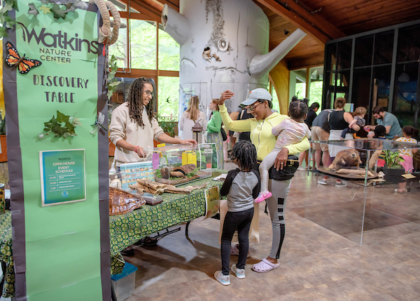 In a large room with a decorative "tree" element and lots of windows, a smiling man behind a table shows items in a plexiglass case to a mother and her kids. A green vertical banner next to the table reads Watkins Nature Center Discovery Table. Behind the mother is a glass case containing a taxidermy beaver.