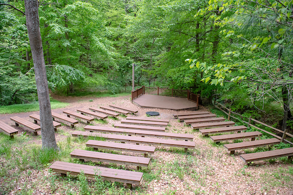 An outdoor amphitheater made of wooden benches sloping down toward a wooden stage platform, all painted brown. The amphitheater is surrounded by green trees.