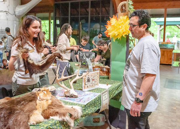 Inside a large room, a smiling woman behind a table holds up a skunk pelt for a man to examine. The table contains other animal hides and antlers, along with information sheets. Behind them is another exhibit table where visitors are looking at something in a plexiglass case.