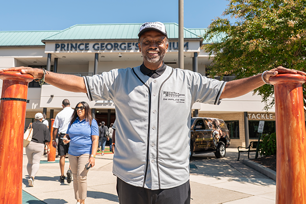Director Stanton standing in front of Prince George's Stadium, wearing a gray baseball jersey with the Parks and Recreation logo. His arms are stretched out to either side, and his hands are resting on two giant baseball bat sculptures.