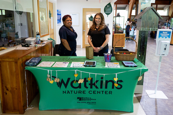 Inside a lobby area, two smiling women stand behind a long table covered with a green tablecloth that reads Watkins Nature Center. The table contains information sheets and a wicker basket, and is decorated with a garland. Behind this area to the right is a larger room with glass terrariums.