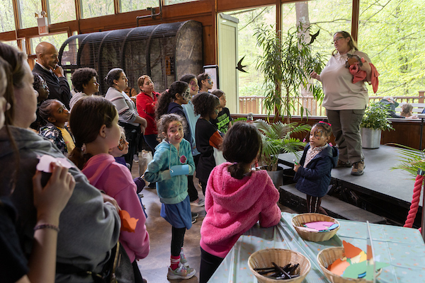 In a large room with a wall of windows, a woman in a staff uniform stands on a platform holding a turtle in her left hand, addressing a rapt audience of children and adults. Several children in the audience have their faces painted.
