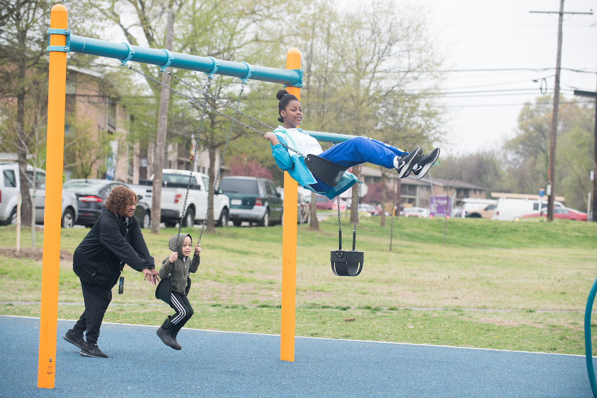 A small child and an older youth play on a swing set at an outdoor playground. The small child is being pushed in his swing by an adult; the older youth is swinging very high on her own.