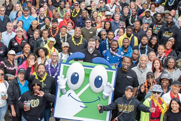 A large group of people huddled together around a big blue and green square character with eyes, hands, and a smile named FUN