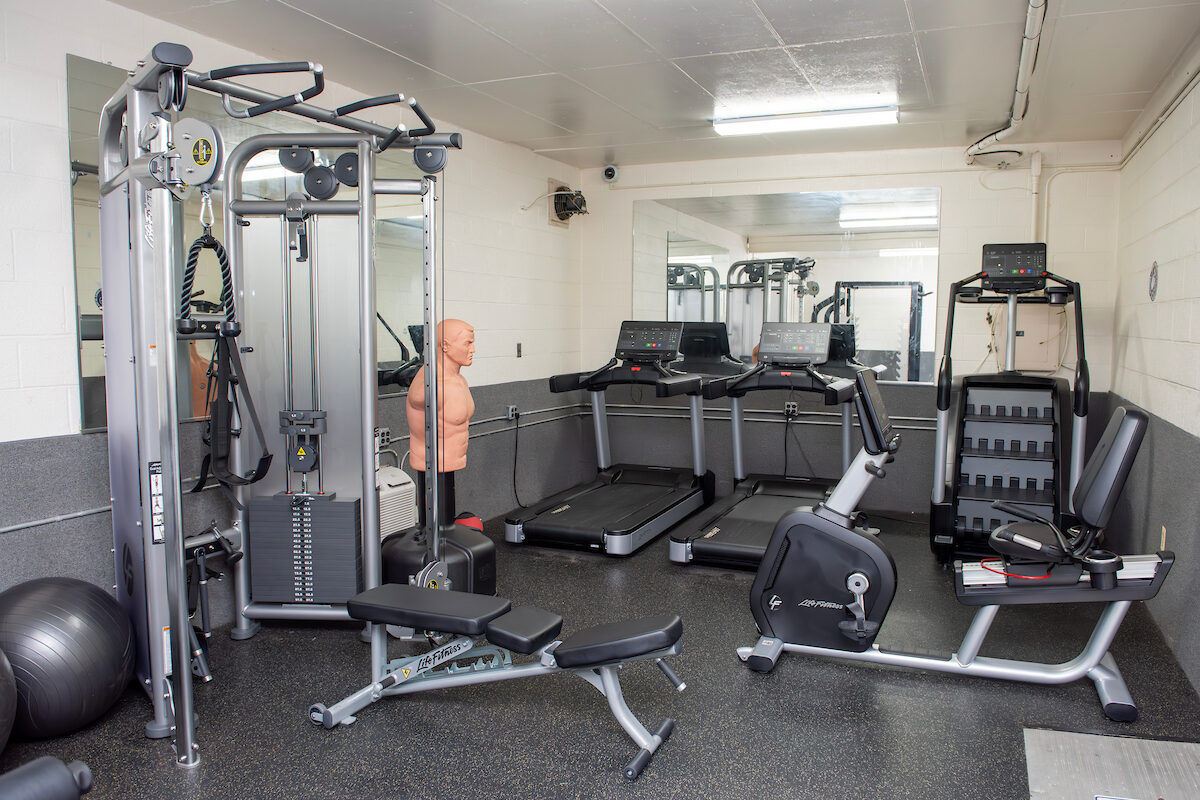 A fitness room with white and gray walls, mirrors on the walls, and multiple pieces of exercise equipment, including treadmills, a stair-climber, a boxing dummy, and a weight machine.