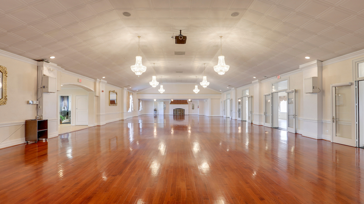 A large ballroom with hardwood floors and six crystal chandeliers hanging from a coffered ceiling. Along the right side are four sets of double doors leading to a sunny adjacent room; in the left side is an arched entryway. There is a fireplace at the far end.