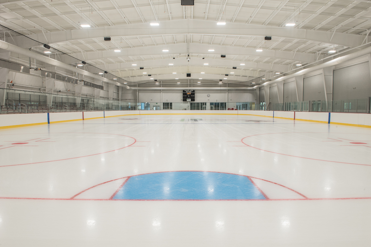 A large ice rink as viewed from one end. The ice is painted with markings for ice hockey, and there is a scoreboard mounted high on the opposite wall.
