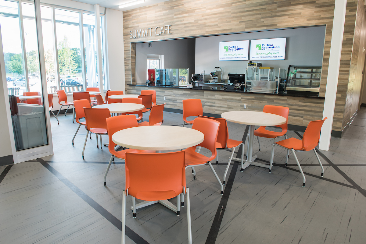 Half a dozen cafe tables, each with three or four orange plastic chairs around it, are arranged in a sunny room in front of a snack bar counter labeled SUMMIT CAFE.