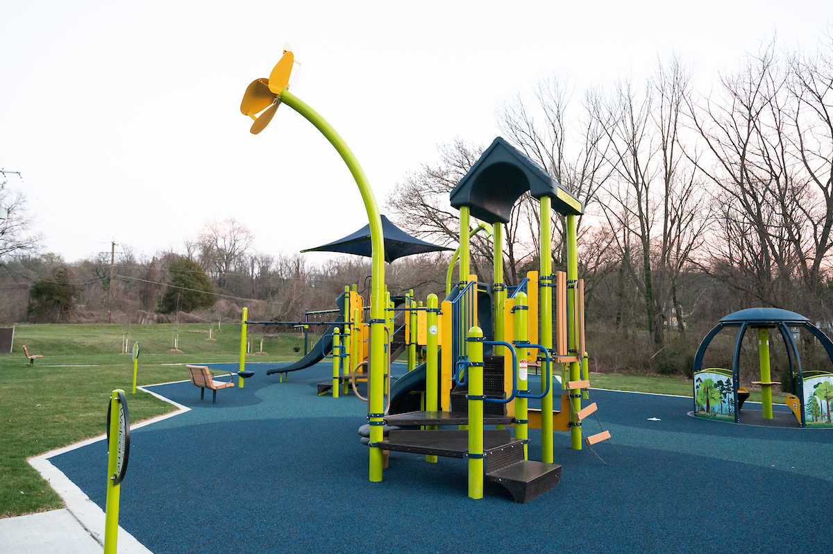 A blue and green outdoor playground featuring several large play structures with platforms, slides, swings, and climbing elements. The green, corner posts of two of the play structures extends up into the sky and become the green stems of giant, whimsical, metal flowers.