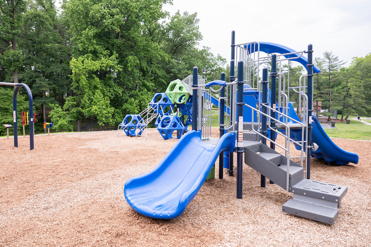 An outdoor playground with several large play structures featuring slides, platforms, stairs, monkey bars, swings, and musical instruments. The playground is blue and green.