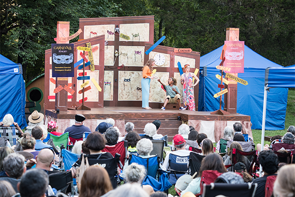 Three actors perform on a small outdoor stage set up in the grass, in front of rows of audience members sitting in lawn chairs. On the stage advertising a carnival, as well as several signposts with arrows labeled with city names including PADUA, LONDON, and PORPENTINE. Behind the stage is a wooded area.