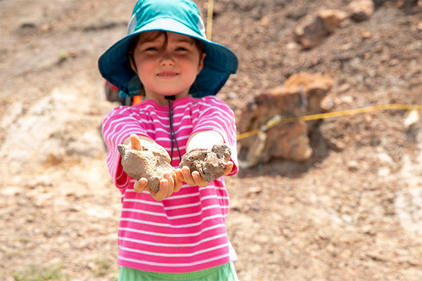 A small child wearing a blue sun hat and a pink shirt smiles while holding two rocky aggregates that might be fossils.