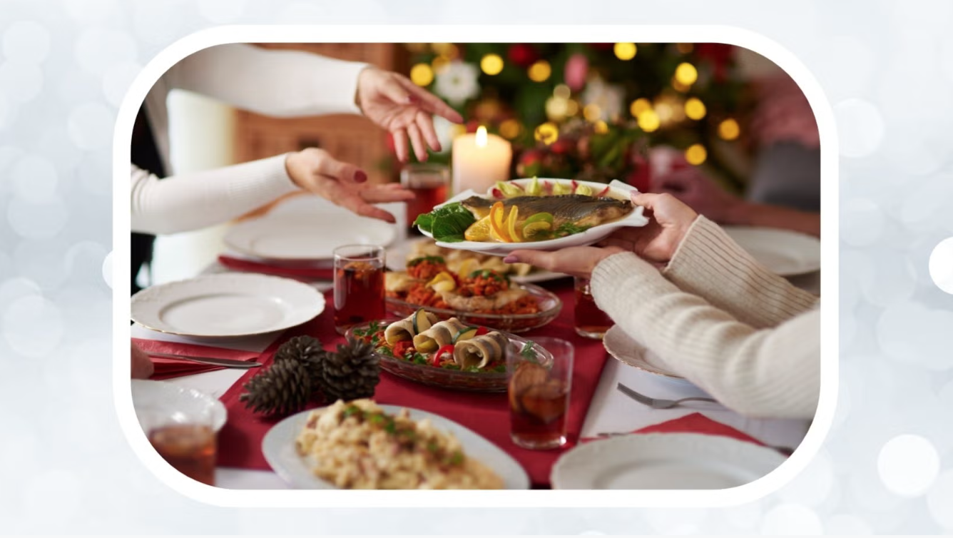 A close-up view of hands passing a plate with a fish on it across a dinner table that is laid with many plates and contains multiple dishes of food. Hands on the other side of the table reach for the fish dish. In the background is a blurry Christmas tree.