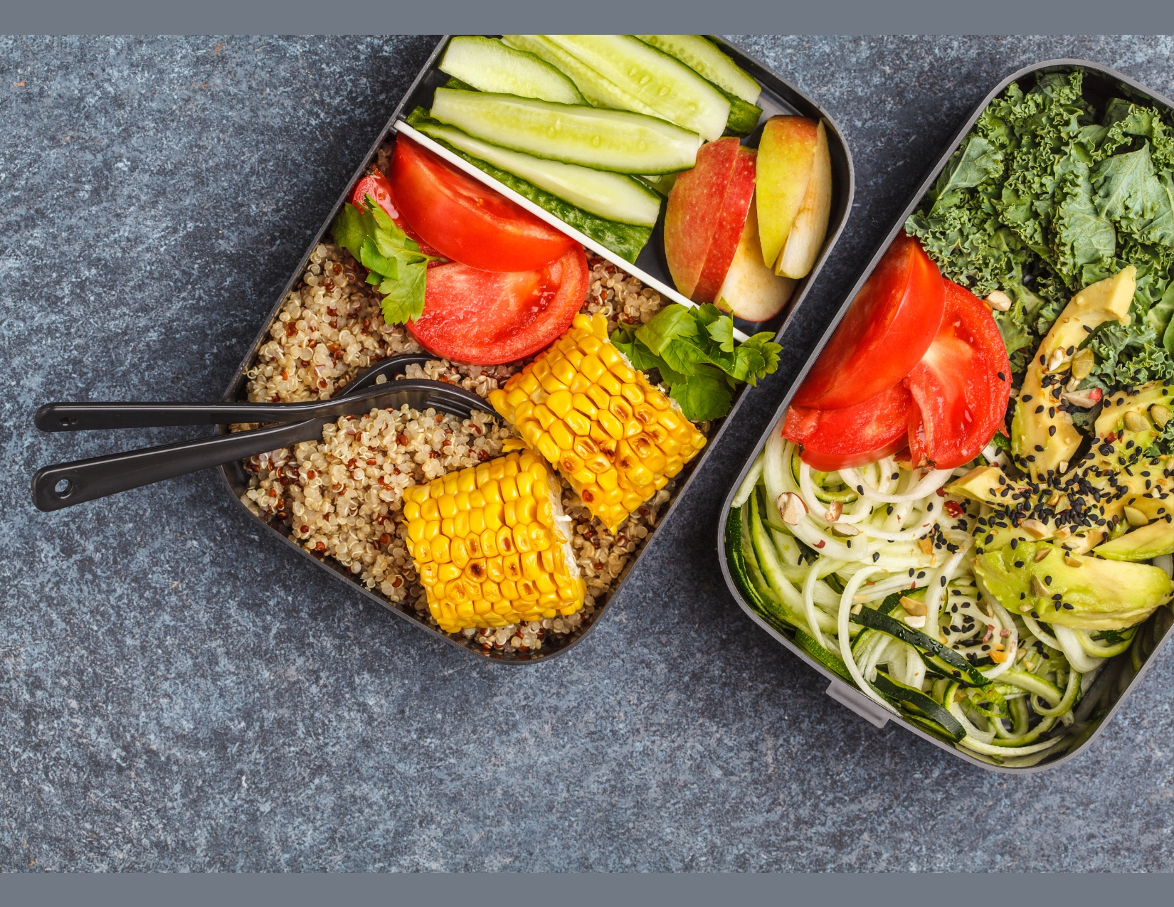Overhead photo of a bento-style lunchbox with several compartments, all containing healthy food: sliced cucumber and apples, quinoa with tomatoes and corn, kale salad with avocado, and spiralized zucchini.