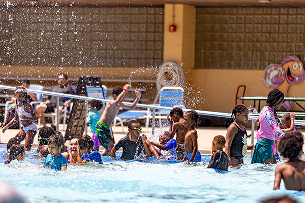Several dozen children play in he shallow end of a pool, splashing water high into the air.