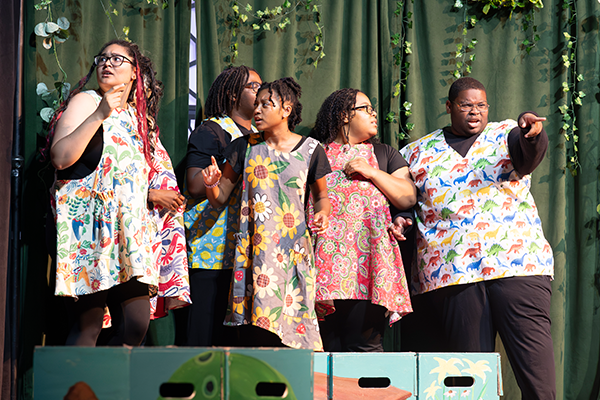 Five young actors wearing printed smocks perform in front of a green backdrop with strands of ivy hanging from it.