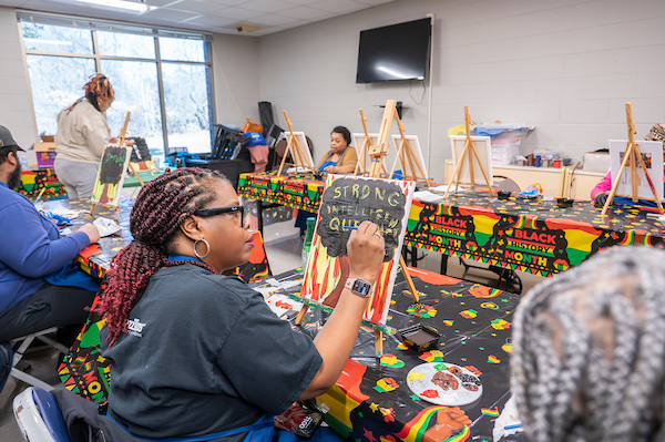 A woman sits at a long table painting on a canvas on a small tabletop easel. Around her are others doing the same. The table has a Black History Month tablecloth on it.