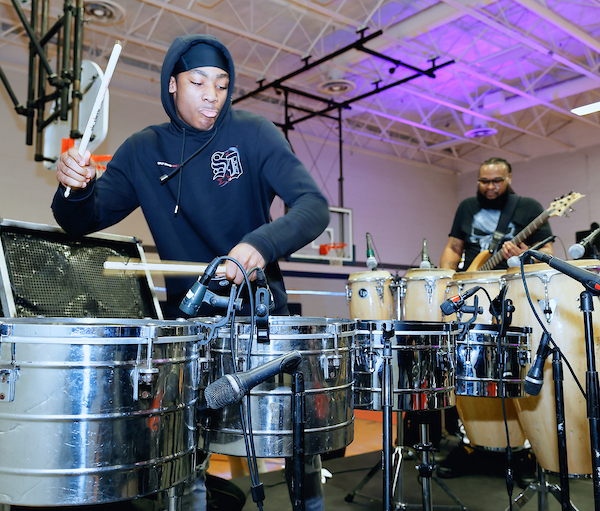 In a gymnasium with purple lighting, a man in a black hoodie plays drums while a man in black t-shirt behind him plays an electric guitar. Between the men is a set of conga drums, all individually mic'd.