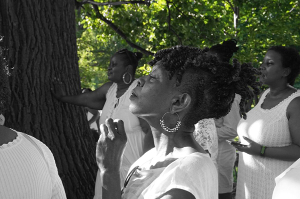 Photographic artwork featuring several women in white dresses depicted in black-and-white against a full-color landscape background of trees with green leaves.
