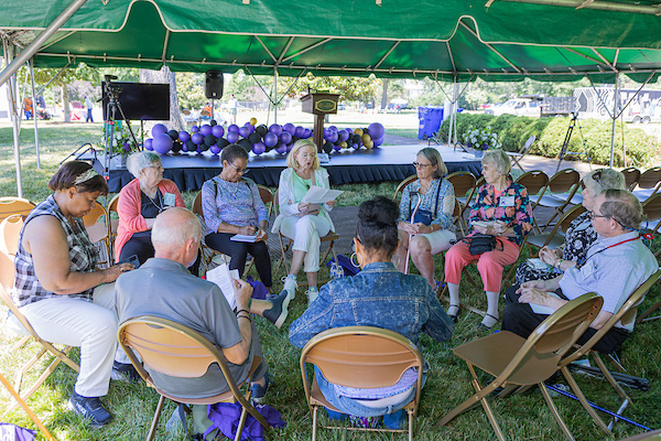 Ten adults sit in a circle of folding chairs in the grass, under a large tent. One is reading aloud from a paper.