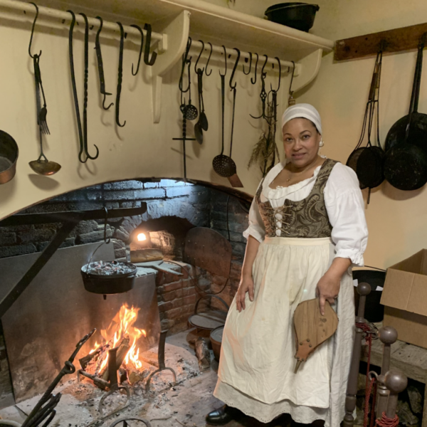 A woman wearing historical colonial clothing standing by a lit brick cooking fireplace holding bellows.