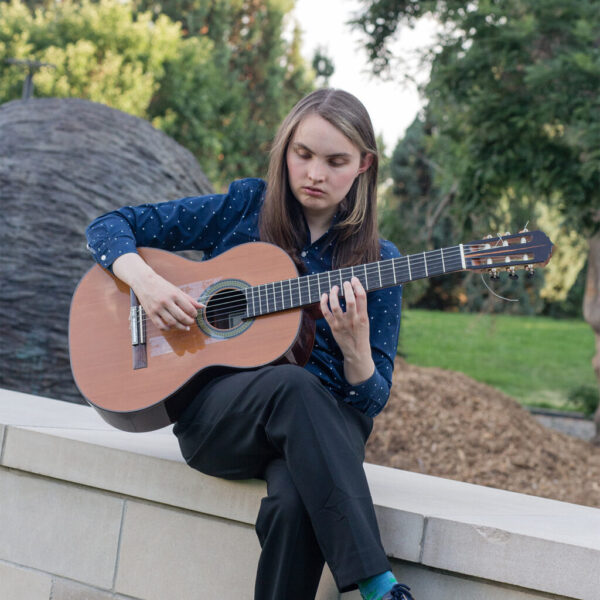 A young woman sitting on a stone wall outdoors playing a classical acoustic guitar.