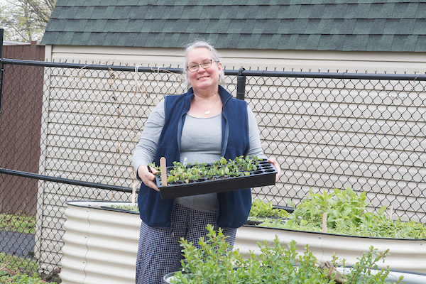 A smiling woman holding a tray of plant seedlings in a garden setting.