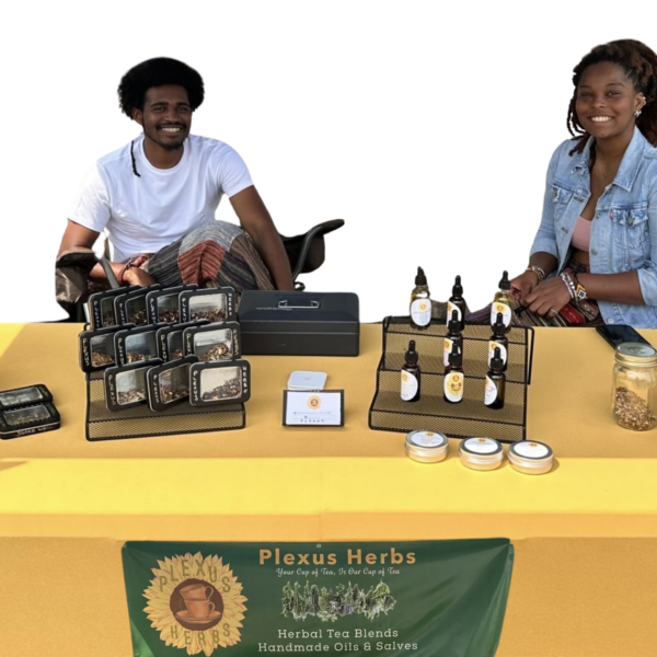 Two vendors smiling behind a yellow table displaying Plexus Herbs teas, salves, and oils.