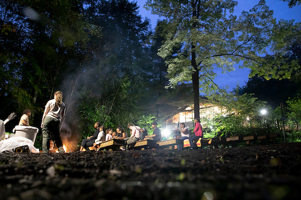 A ranger speaks to people sitting in an amphitheater at dusk. There is a campfire between the audience and the ranger.