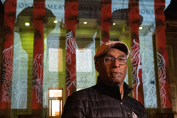 A man in a black jacket poses in front of a projection on the exterior of a building at night.