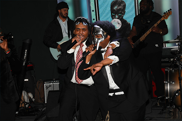 Two men in suits perform with handheld microphones onstage, with a backing band behind them. One wears a silver, MF Doom-style mask.